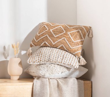 Elevating a warm, bohemian-inspired nook, stack of textured decorative pillows with intricate woven jute tassels sits atop a light wood bench beside a small ceramic vase, minimalist interior scheme. clipart