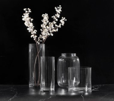 Elegant clear glass vases in varying shapes, styled with delicate white blossoms, set against a dramatic black backdrop, highlighting modern and minimalist interior decor concepts.