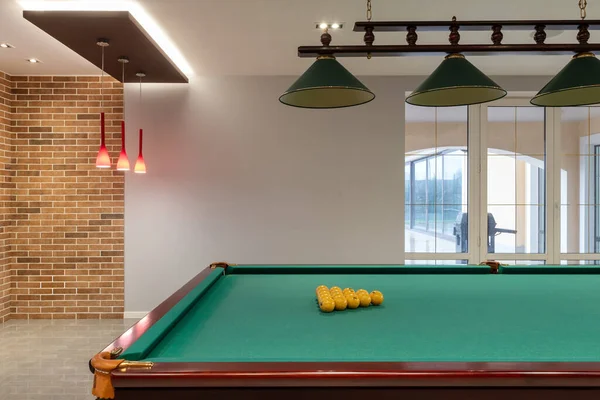 A billiard table with yellow balls sits in a billiard room with a fireplace, wooden walls, and a large window overlooking the property
