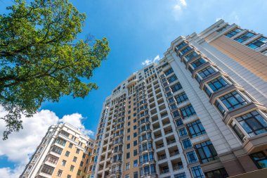 Modern apartman binaları ve ağaçlar mavi gökyüzüne karşı kentsel yaşam ve mimariyi gösteriyor.