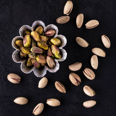 Üst görünüm siyah desenli arka plan, kabuklu fıstık tohumlu kâse; arka planda kabuklu fıstıklar.