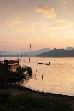 Günbatımında Mekong Nehri manzarası, nehir kıyısında geleneksel Laos feribot iskelesi ve nehirde kürek çeken bir balıkçı. Luang Prabang, Laos.