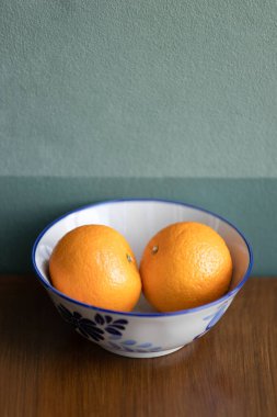 Kahverengi meşe ağacının üzerinde beyaz bir seramik kasede iki portakal meyvesi, arka planda koyu yeşil duvar..