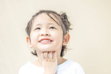 Asyalı anaokulu kızı uzun kollu beyaz bir tişört giyip kollarını çapraz yapıyor, elleri çenesinde, yukarı bakıyor ve gülümsüyor..