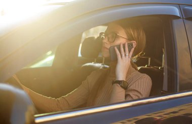 Genç iş kadını araba kullanıyor, akıllı bir telefonla konuşuyor, gün batımının arka planına karşı.