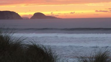 İki kız, okyanusta gün batımının arka planında sörf yapıyor. Uzakta kıyı kayaları silueti ve ön planda kıyı çimenleri olan kumlu bir plaj var.
