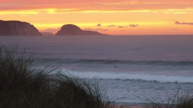 İki kız, okyanusta gün batımının arka planında sörf yapıyor. Uzakta kıyı kayaları silueti ve ön planda kıyı çimenleri olan kumlu bir plaj var.