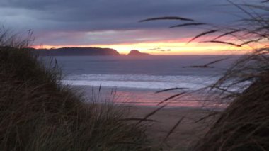 Okyanusta harika bir gün batımı. Uzaktaki sahil kayaları silueti ve ön planda kıyı çimenleri olan kumlu bir sahil. Arka planda kayalıklar.