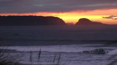 Okyanusta harika bir gün batımı. Uzaktaki sahil kayaları silueti ve ön planda kıyı otları olan kumlu bir sahil.