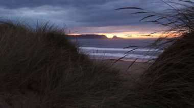 Okyanusta harika bir gün batımı. Uzaktaki sahil kayaları silueti ve ön planda kıyı otları olan kumlu bir sahil.