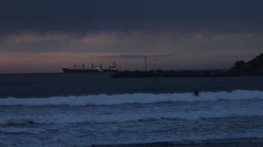 Akşamları birkaç kişi, ışıkları açık ve rıhtımı olan bir tankerin arka planına karşı okyanusta sörf yapıyor.