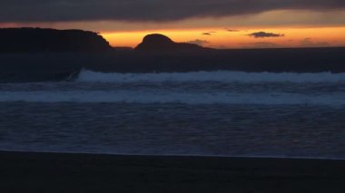 Okyanusta harika bir gün batımı. Uzakta kıyı kayaları silueti ve ön planda kumlu bir plaj var.