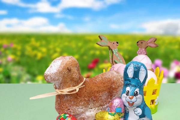 Easter card. A traditional Easter lamb cake, a easter bunny, chocolate sweets and basket with eggs and wooden bunnies over abstract blurred spring background.