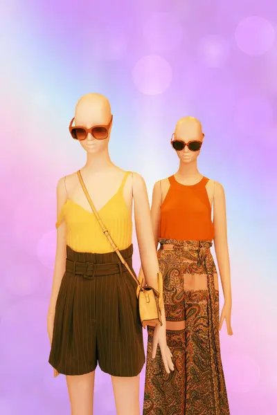 Two mannequins dressed with yellow shirt, elegant brown shorts and handbag, sunglasses and the other in orange shirt with long skirt over abstract summer background. Space.
