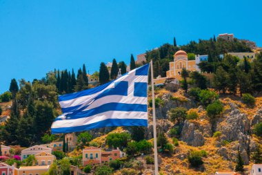 SYMI, Yunanistan - Haziran 03, 2021 Sarp dağ yamaçlarında yükselen renkli villaların önünde bir Yunan bayrağı sallıyor.