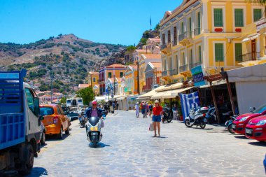 SYMI, Yunanistan - 3 Haziran 2021. Symi Town limanındaki turistler. Renkli villalar dik yamaçlara uzanıyor..