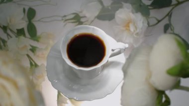 Güneş ışığıyla aydınlatılan çiçeklerin arka planında bir fincan kahve.
