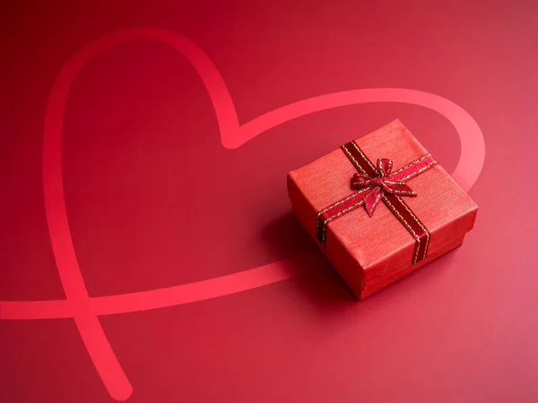 A small red present gift box with ribbon on heart shape drawing red background with copy space, top view. An essential gift on special days, birthdays, New Years, Valentine's Day and anniversaries.