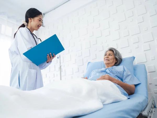 stock image Smile Asian senior woman grey hair patient lying on bed, recovering while young female doctors visit, explaining the symptoms and examination notes and medical healthcare documents in hospital room. 