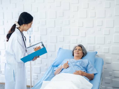 Sırıtkan Asyalı son sınıf öğrencisi gri saçlı hasta, genç bayan doktorlar geldiğinde iyileşiyor, hastane odasındaki semptomları, muayene notlarını ve tıbbi sağlık raporlarını açıklıyor.. 