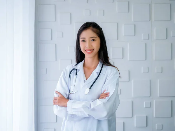 Smiling pretty Asian woman doctor portrait long hair, standing with arms cross in white modern room at hospital. Confident Asian young female practitioner with stethoscope, looking at camera.