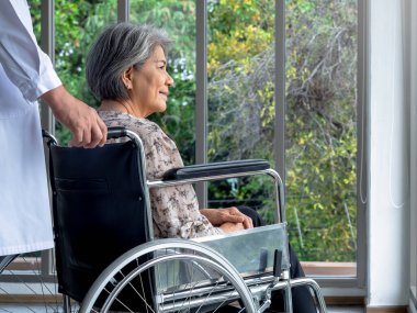 Mutlu Asyalı yaşlı kadın hastanın yakın plan portresi tekerlekli sandalyede otururken beyaz önlüklü adam hastanedeki muayenehanede tedavi görüyor. Bakım, sağlık ve tıbbi konsept.