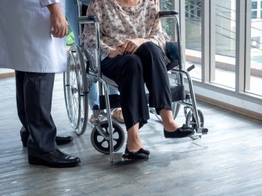 Yaşlı hasta kadın tekerlekli sandalyede yerde otururken beyaz önlüklü adam hastanede bekleme odasında onu ziyaret etmek için yanında duruyor. Bakım, sağlık ve tıbbi konsept.