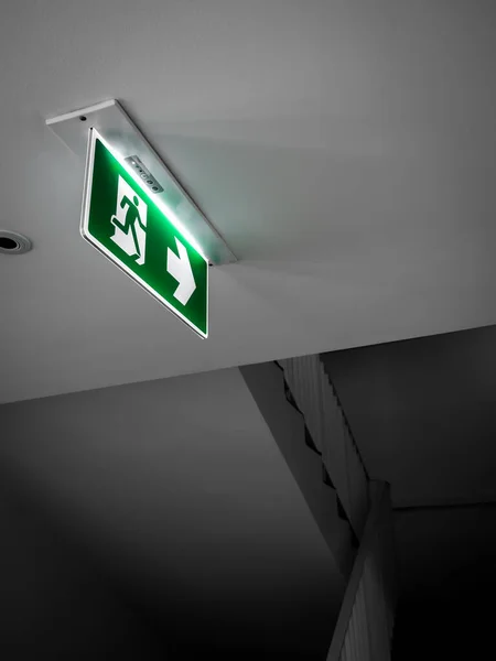 Fire exit sign. Green emergency exit sign hanging from white ceiling, glowing at the dark corridor near the stairway fire escape door in the building, vertical style.