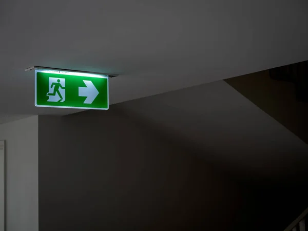 Green fire escape sign hang on the ceiling in the dark building near the stairway. Emergency fire exit sign, warning plate with running man icon and arrow to the right way with copy space.