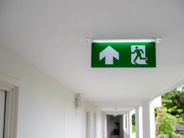 Green fire escape sign hang on the ceiling at the corridor in the white hotel building near the stairway. Emergency fire exit sign, warning plate with running man icon and arrow to the straight way.