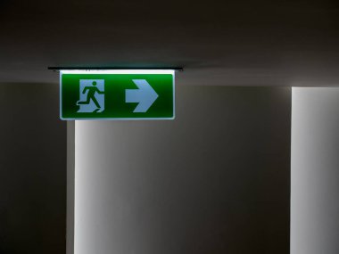 Green fire escape sign hang on the ceiling in the dark building. Emergency fire exit sign, warning plate with running man icon and arrow to the right way with copy space.