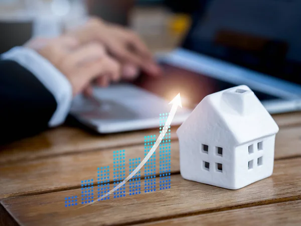 Property value, real estate investment, mortgage and home tax concepts. Glow rising up arrow on digital business chart near white miniature house and business person working with laptop computer.