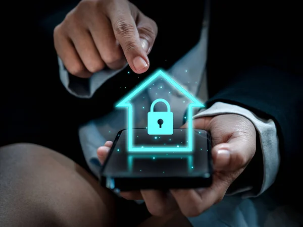Smart home technology concept. Glowing digital security home lock symbol hologram appearing on smartphone in businessperson's hands. Smarthome safety system automation control app.