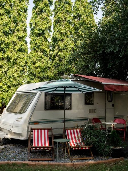 Vintage caravan car parking in garden decoration with empty seats and table on big tree background, vertical style. Relax camping and sleep in the motorhome trailer. Family vacation travel concept.