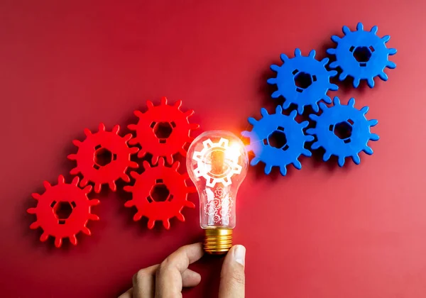 Digital gear wheel symbol appear on light bulb in businessman\'s hand with blue and red gears on red background. Business strategy, management and solution, human research, teamwork and idea concepts.