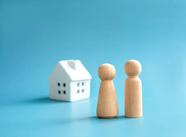 Love couple wooden figure model, male and female symbol, standing front of small white house miniature on blue background. Building family. First home, buying house, real estate investment concept.