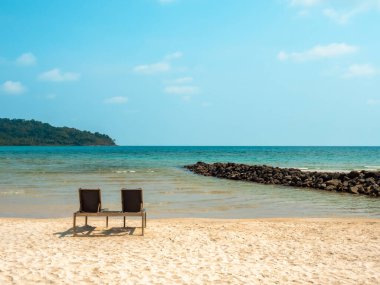 Adanın kayalıklarının yanındaki mavi denizin önündeki kumlu sahilde boş güneşli yatak koltukları. Güneşli bir günde deniz manzaralı arka planda iki sandalye dinleniyor..