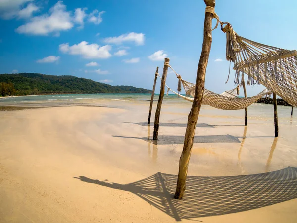 stock image Three empty rope cradle hanging on the wooden poles on sandy beach, island and blue sky background, fisheye view. Beautiful shadow of hanging cradle or swing beds on clean sand beach.