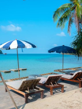Mavi çizgili ayakta duran plaj şemsiyeli, mavi deniz manzaralı ve yaz gökyüzü arka planlı, iki tahta güneşli yatak, dikey. Tatil gezisi geçmişi.
