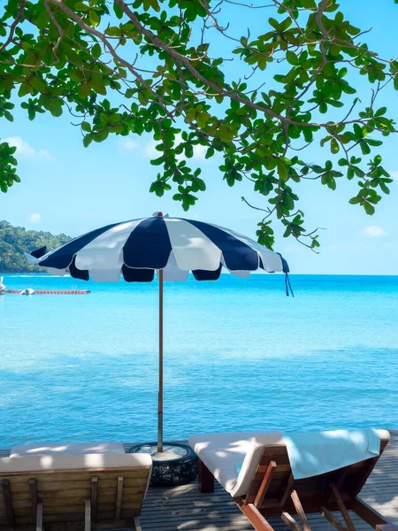 两张木制的日光浴床 床垫装饰着蓝色条纹的立式海滩伞 海景上有蓝色的大海和开阔的天空背景 垂直风格 假日背景 — 图库照片
