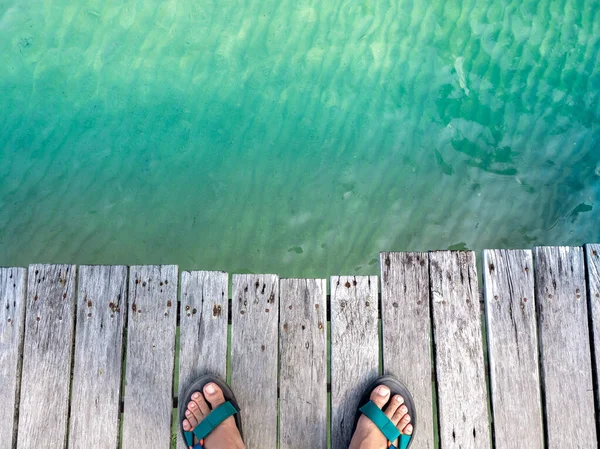在岛上海滩附近 穿着凉鞋站在干净清澈的海水之上的旧木桥上的人的脚的头像 有复制的空间 暑假海报背景 — 图库照片