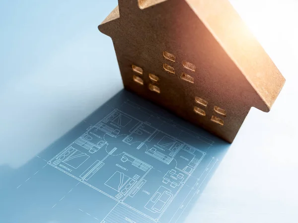 Home floor plan in the shadow near the small wooden house model on blue background. Concepts of home building, decoration, interior design, business success and goal idea and inspiration.