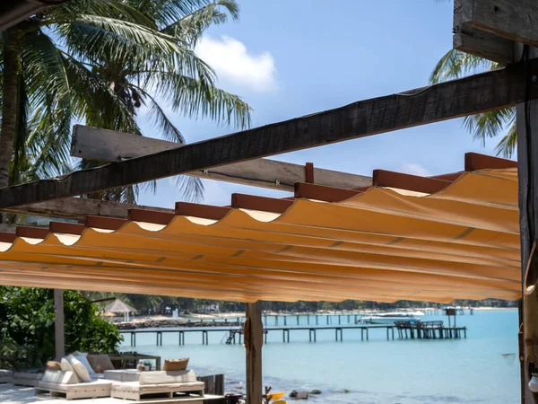 Outdoor retractable roof on wood and iron construction on the beach front restaurant near the tropical palm tree and seascape view background. Foldable fabric canvas awning on the seaside.