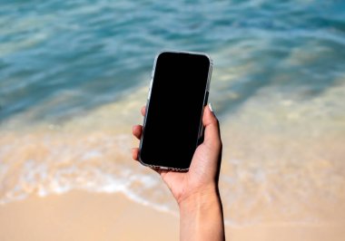 Boş ekranlı cep telefonu. Yazın mavi deniz arka planında elinde cep telefonu tutan siyah boş masaüstü ekran görüntüsü. Seyahat rezervasyonu, arama, iş pazarlaması.
