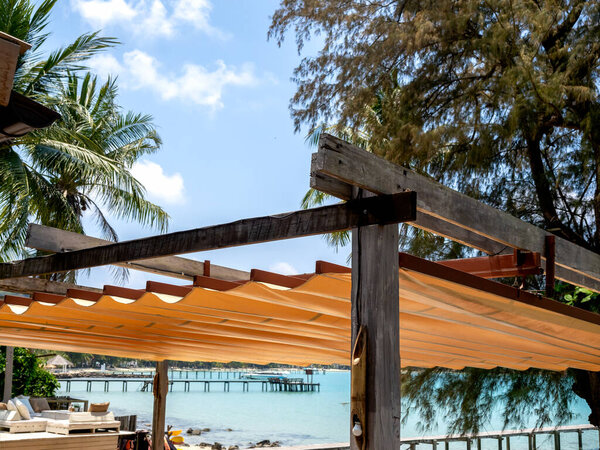 Outdoor retractable roof on wood and iron construction on the beach front restaurant near the tropical palm tree and seascape view background. Foldable fabric canvas awning on the seaside.