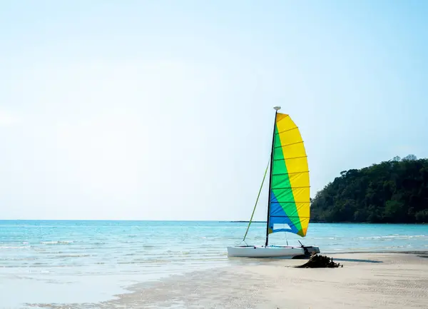 在阳光明媚的夏日 一艘装着黄色 蓝色和绿色图案帆的空白色帆船停泊在沙滩上 在大海 岛屿和蓝天的衬托下 显得格外引人注目 — 图库照片