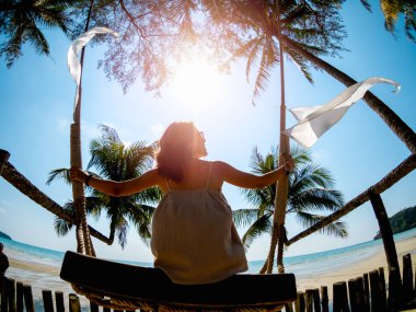 Asyalı bir kadının arkası günlük kıyafetlerle yaz aylarında plajın önünde güneş ışığıyla salıncakta oturuyor. Yalnız kadın denizde dinleniyor ve gökyüzüne bakıyor, deniz manzarası, tatil tatili.