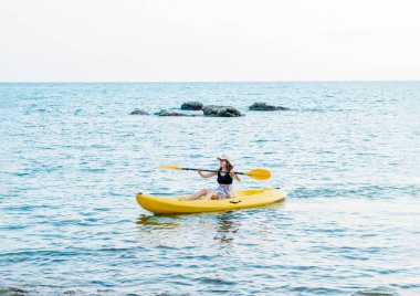 Siyah kolsuz tişörtlü, sarı kano kayığında kano süren ve denizde kürek çeken mutlu Asyalı kadın. Mutlu kadın deniz manzaralı arka planda eğlenceli aktiviteler, tatil gezisi.