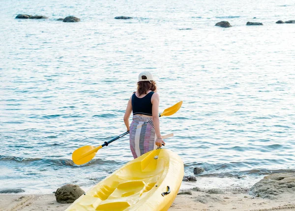 Asian woman in black tank top and cap dragging yellow kayak and holding paddle on the sand to the sea. Happy female having fun activity at the beach front blue sky background, Holiday trip vacation.