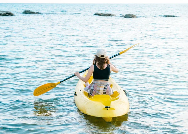Back of Asian woman in black tank top and cap kayaking on yellow kayak boat with using paddle on the sea. Happy female having fun activity on seascape view background, Holiday trip vacation.